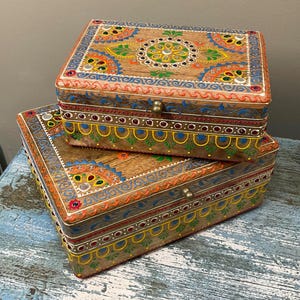 May include: Two wooden decorative boxes with detailed hand-painted designs. The boxes have a natural wood grain base, decorated with colourful floral and geometric patterns in orange, blue, green, and yellow. Each box has a brass clasp.