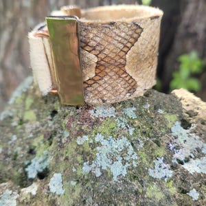 May include: A wide cuff bracelet featuring a textured snakeskin pattern in shades of tan and brown. The bracelet includes a smooth, polished gold-tone metal rectangular clasp and a section of off-white textured material on the side.