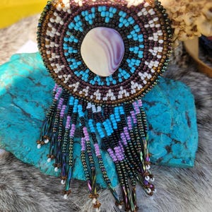 May include: A handcrafted beaded brooch with a central oval stone. The brooch features a circular design with turquoise, purple, white, and brown beads. Long beaded fringe in shades of purple, black, and iridescent green hangs below the brooch.