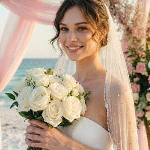 May include: A bride smiles, holding a bouquet of white roses. She wears a strapless white dress and a veil with pearl accents. The background includes a beach, pink draping, and floral arrangements, suggesting a wedding ceremony.