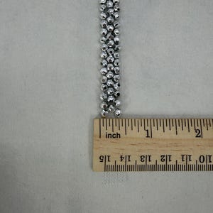 May include: A silver beaded strand is measured with a wooden ruler. The strand is made of small, round, metallic beads. The ruler shows the strand is approximately 1.5 inches long.
