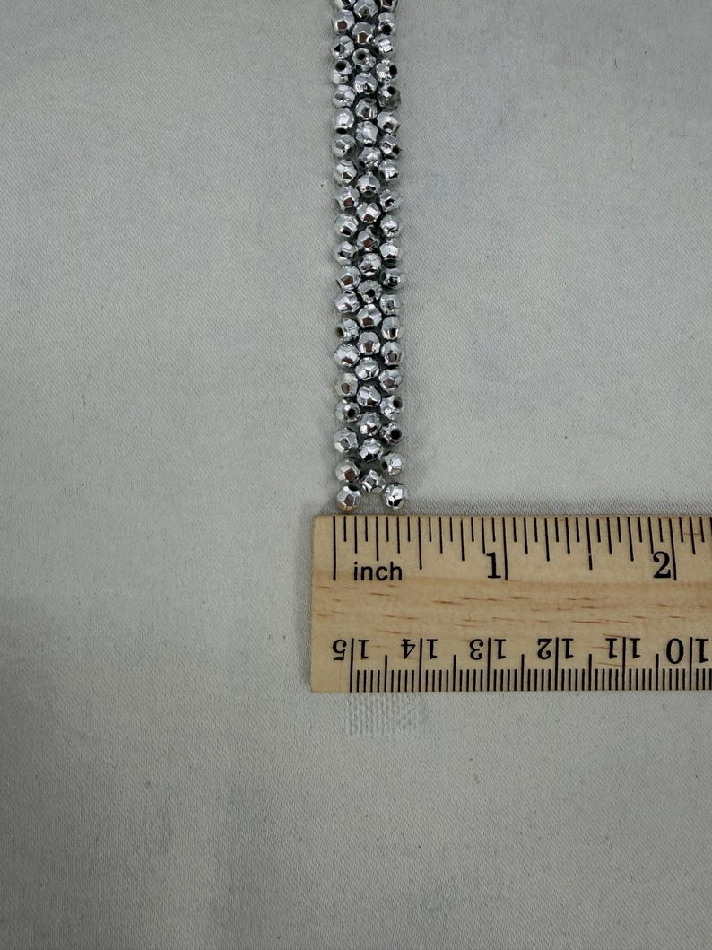 May include: A silver beaded strand is measured with a wooden ruler. The strand is made of small, round, metallic beads. The ruler shows the strand is approximately 1.5 inches long.