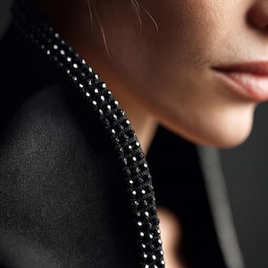 May include: Close-up of a black jacket collar with a beaded trim. The trim is made of small, white, reflective beads. The jacket is made of a smooth, dark fabric. The background is dark and out of focus.