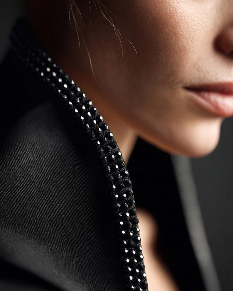 May include: Close-up of a black jacket collar with a beaded trim. The trim is made of small, white, reflective beads. The jacket is made of a smooth, dark fabric. The background is dark and out of focus.