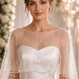 May include: A sheer white tulle bridal capelet featuring a delicate scalloped edge trimmed with small white pearls and scattered pearl embellishments across the fabric, worn over a strapless white satin bodice with a sweetheart neckline.