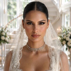 May include: A bride wearing a white wedding dress with a beaded veil and a multi-strand diamond choker necklace. The bride has dark hair pulled back and is wearing makeup. The background includes a staircase and floral arrangements.