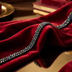 May include: A close-up shot of a deep red velvet fabric with a beaded strand. The beads are iridescent, reflecting multiple colors. The fabric is draped, creating soft folds and shadows. The overall aesthetic is luxurious and elegant.