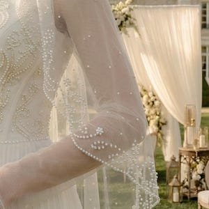 May include: A white wedding dress with a sheer, long-sleeved veil. The veil and dress are adorned with small, delicate pearls. The dress has intricate embroidery on the bodice. The background features a garden setting with floral decorations.
