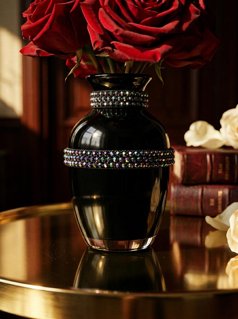 May include: A black vase with two bands of iridescent jewels, holding a bouquet of deep red roses. The vase sits on a gold-toned surface, with a blurred background of books and flowers.