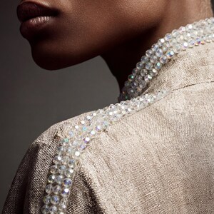 May include: Close-up of a tan-colored jacket with a beaded embellishment along the shoulder line. The beads are clear and iridescent, adding a touch of sparkle. The jacket has a textured appearance, suggesting a woven fabric.