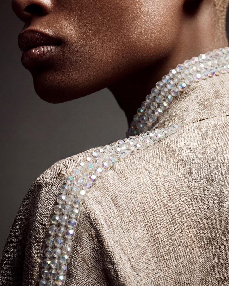 May include: Close-up of a tan-colored jacket with a beaded embellishment along the shoulder line. The beads are clear and iridescent, adding a touch of sparkle. The jacket has a textured appearance, suggesting a woven fabric.