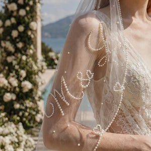 May include: A sheer white wedding veil adorned with delicate pearl embellishments. The veil features the word "Mrs" spelled out in pearls on the sleeve, with heart-shaped pearl accents. The veil is part of a bridal ensemble.