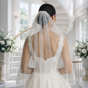 May include: A bride in a white wedding dress and veil, viewed from the back. The dress features intricate lace detailing and a full skirt. The veil is adorned with small, scattered pearls. The background includes a grand staircase and floral arrangements.