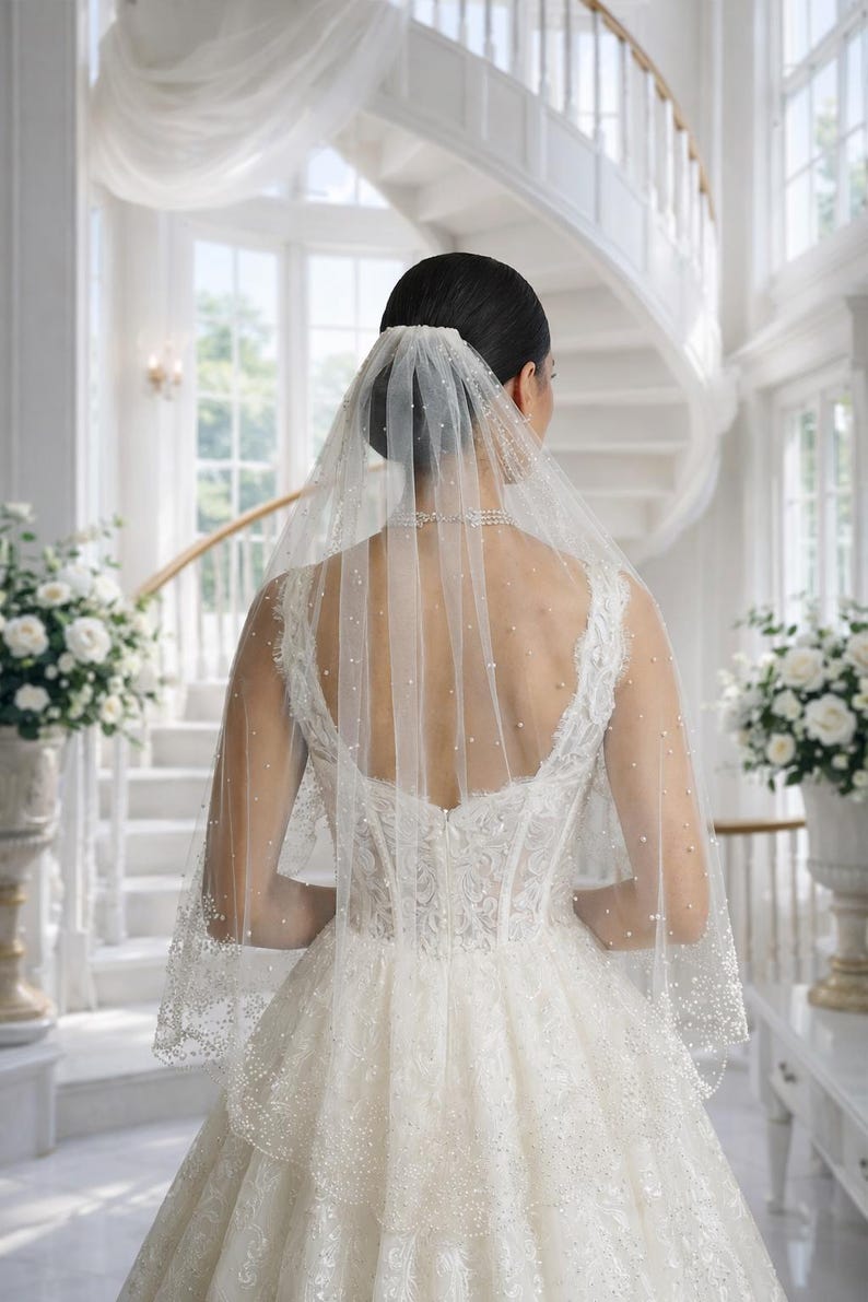 May include: A bride in a white wedding dress and veil, viewed from the back. The dress features intricate lace detailing and a full skirt. The veil is adorned with small, scattered pearls. The background includes a grand staircase and floral arrangements.