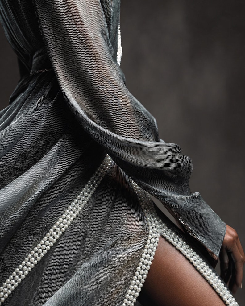 May include: A flowing, silver-grey dress with a high slit, trimmed with rows of small, white pearls. The dress has long sleeves and a sheer, shimmering fabric, creating an elegant and sophisticated look. The dress is perfect for a special occasion.