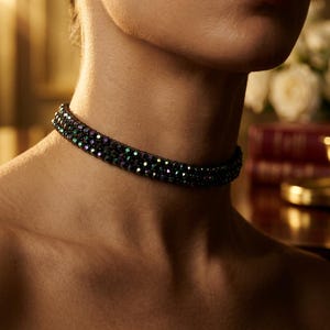 May include: A close-up of a black beaded choker necklace with iridescent green and purple accents. The necklace is worn around the neck. The background is blurred, with soft lighting.