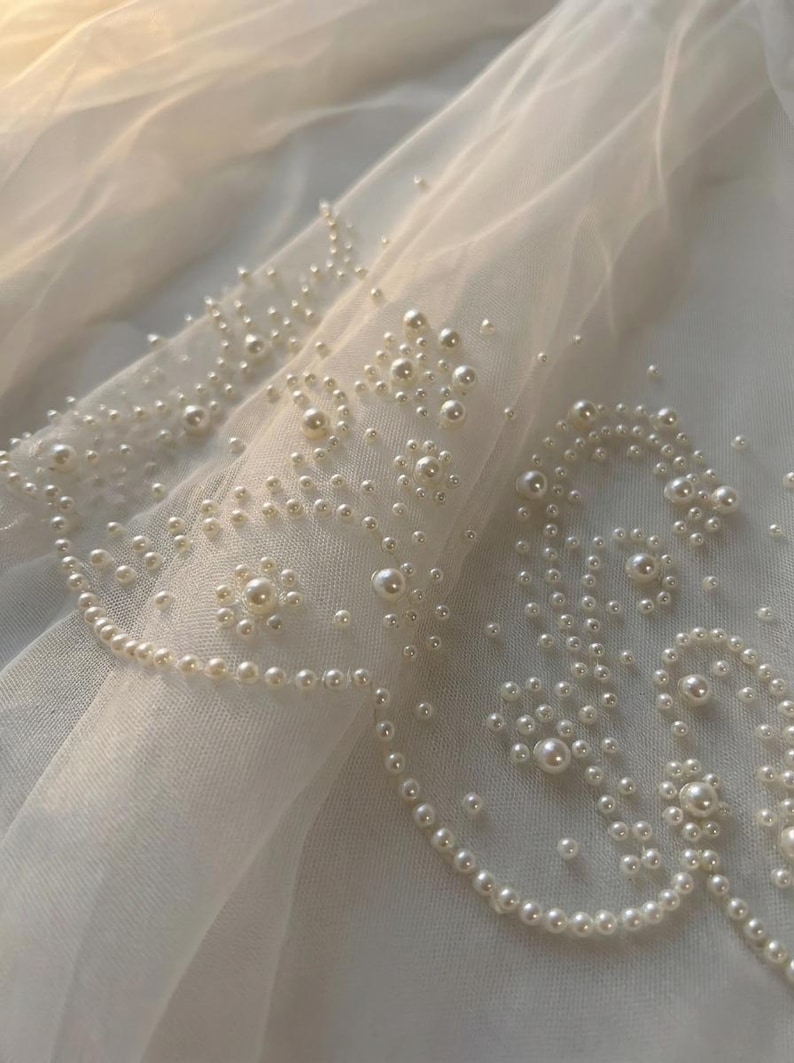 May include: Close-up of a sheer, white fabric adorned with numerous, small, off-white pearls. The pearls are arranged in floral and curved patterns, creating a delicate, textured design. The fabric is softly lit, highlighting the pearl embellishments.