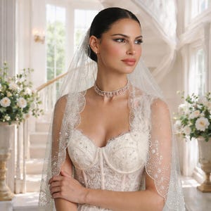 May include: A bride in an ivory wedding dress with a sweetheart neckline and intricate lace detailing. She wears a sheer veil adorned with delicate embellishments and a matching choker necklace. The setting is a bright, elegant space with a staircase and floral arrangements.