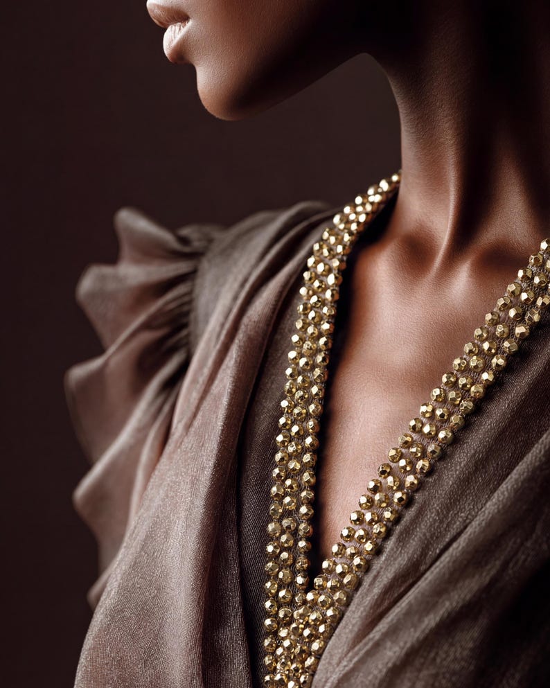 May include: A close-up of a person wearing a brown dress and a gold beaded necklace. The necklace is made of small, faceted beads and drapes down the neckline. The dress has a flowing, draped design.