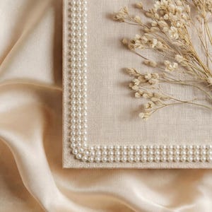 May include: A rectangular, beige-colored box with a pearl border. Dried, light-brown flowers rest on the box. The background is a cream-colored, silky fabric, creating a soft, elegant aesthetic.