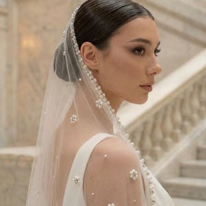 May include: A sheer white bridal veil with pearl and floral embellishments. The veil is adorned with small pearls and larger floral accents, creating a delicate and elegant look. The veil is draped over a white dress.