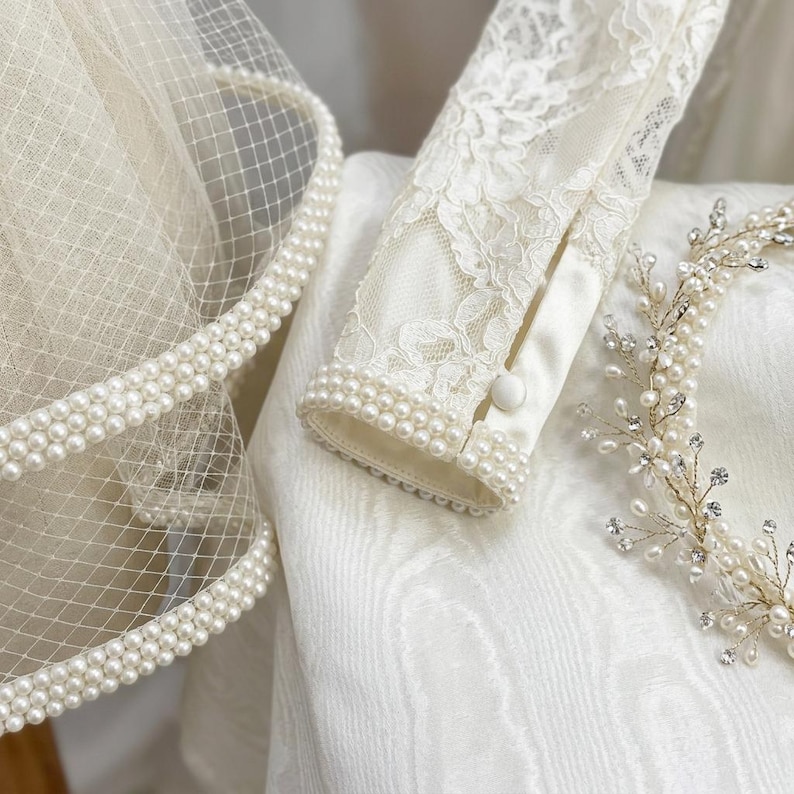 May include: Ivory-colored bridal ensemble featuring a veil with a pearl-beaded edge, a lace-sleeved top with pearl cuff detailing, and a delicate pearl and crystal headpiece. The items are arranged on a white textured surface.