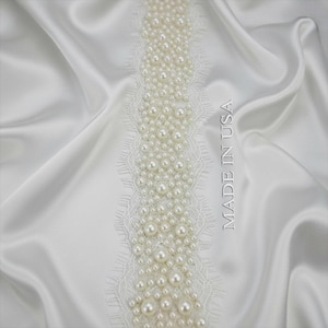 May include: A white lace trim embellished with numerous round, glossy white pearls of varying sizes, arranged in a dense vertical pattern. The trim is set against a smooth, draped white satin fabric. The text MADE IN USA is visible.