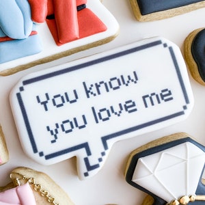 Puede incluir: Galletas de azúcar decoradas con varios diseños. Una galleta tiene la forma de un bocadillo con el texto "You know you love me". Otras galletas tienen forma de bolso, anillo de diamantes y persona.
