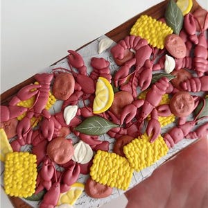 May include: A decorative wooden tray filled with crawfish, corn, lemon slices, and garlic cloves. The crawfish are a reddish-pink color, and the corn is yellow. The tray is held by a hand.