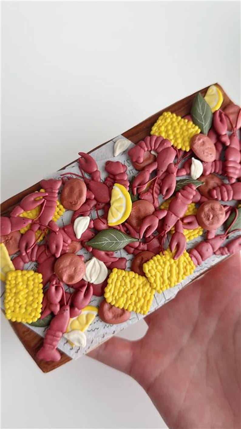 May include: A decorative wooden tray filled with crawfish, corn, lemon slices, and garlic cloves. The crawfish are a reddish-pink color, and the corn is yellow. The tray is held by a hand.