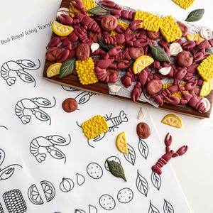 May include: A wooden serving board is covered with edible crawfish, corn, lemon slices, and other food decorations. The board is surrounded by loose decorations and a sheet of paper with the text "Boil Royal Icing Transfer" and crawfish illustrations.