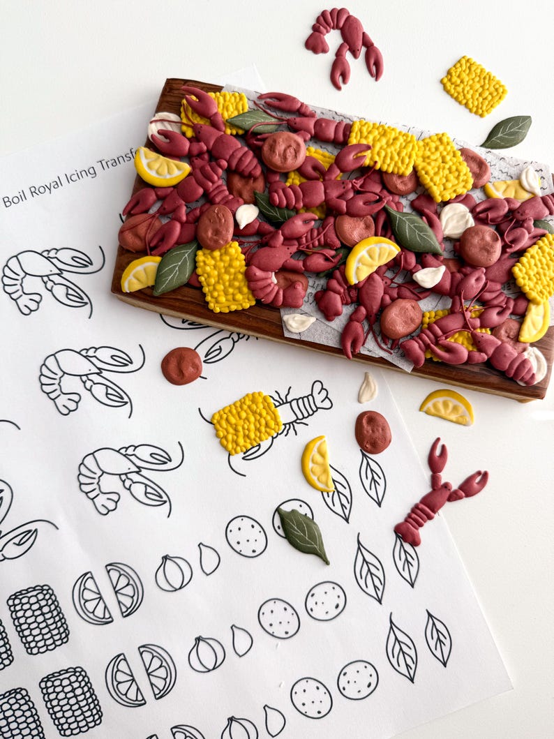 May include: A wooden serving board is covered with edible crawfish, corn, lemon slices, and other food decorations. The board is surrounded by loose decorations and a sheet of paper with the text "Boil Royal Icing Transfer" and crawfish illustrations.