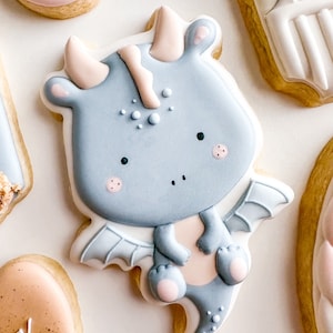 Puede incluir: Una galleta azul claro decorada con un diseño de dragón. El dragón tiene alas rosas, una nariz rosa y puntos blancos. La galleta es de color marrón claro.