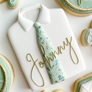 Peut inclure: Un biscuit en sucre blanc décoré pour ressembler à une chemise habillée avec une cravate florale verte et blanche. Le nom "Johnny" est écrit en cursive dorée sur le biscuit.