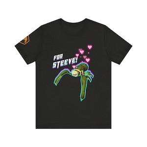 May include: Dark grey t-shirt with a green and tan creature graphic and the text "FOR STEEVE!" in a retro style. Pink hearts float above the creature. An emblem in gold and orange is on the left sleeve.