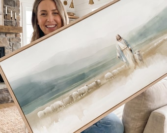 May include: A rectangular landscape painting in a light wood frame depicting a figure in a white and blue robe walking through a field with a flock of sheep. The background features soft, muted green and beige hills under a hazy, light sky.