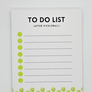 Op de afbeelding: Wit notitieblok met zwarte tekst die "TO DO LIST (AFTER PICKLEBALL)" luidt. Het notitieblok heeft bovenaan elke regel een groene pickleball-afbeelding en een rand van groene pickleball-afbeeldingen onderaan.