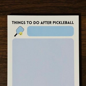 Op de afbeelding: Een wit notitieblok met de tekst "THINGS TO DO AFTER PICKLEBALL" in zwart. Bovenaan staat een blauwe rechthoek met een gele smiley-gezicht pickleball-bat. Onderaan staan drie gele smiley-gezicht pickleball-ballen.
