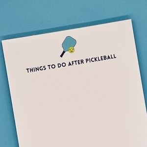 Op de afbeelding: Een wit notitieblok met de tekst "THINGS TO DO AFTER PICKLEBALL" en een afbeelding van een blauwe paddle en een gele bal.