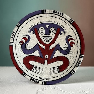 May include: A round ceramic plate featuring a speckled white background with a central stylized figure in red and purple. The figure has large eyes, a wide grin, and raised arms. The rim is decorated with black dashes, and the word PANAMA is printed.