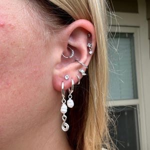Puede incluir: Una curaci&oacute;n de oreja en plata con m&uacute;ltiples piercings. El l&oacute;bulo muestra dos pendientes de aro plateados, uno con una perla blanca colgante y un dije de alambre en espiral, el otro con una perla sencilla. Otros piercings incluyen un aro daith, un pendiente plano y varios aros y tachuelas plateadas peque&ntilde;as a lo largo del cart&iacute;lago.