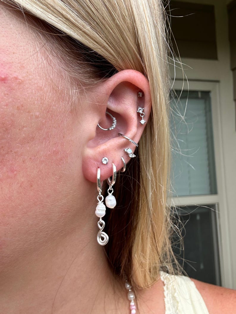 Puede incluir: Una curaci&oacute;n de oreja en plata con m&uacute;ltiples piercings. El l&oacute;bulo muestra dos pendientes de aro plateados, uno con una perla blanca colgante y un dije de alambre en espiral, el otro con una perla sencilla. Otros piercings incluyen un aro daith, un pendiente plano y varios aros y tachuelas plateadas peque&ntilde;as a lo largo del cart&iacute;lago.