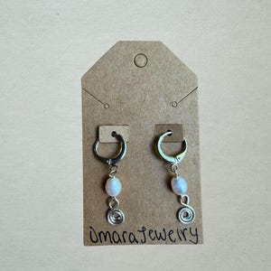 Puede incluir: Un par de pendientes de aro plateados con una cuenta de perla ovalada blanca y un dije de espiral de alambre plateado. Los pendientes est&aacute;n sujetos a una tarjeta de presentaci&oacute;n de cart&oacute;n marr&oacute;n con el texto Omara Jewelry escrito a mano.