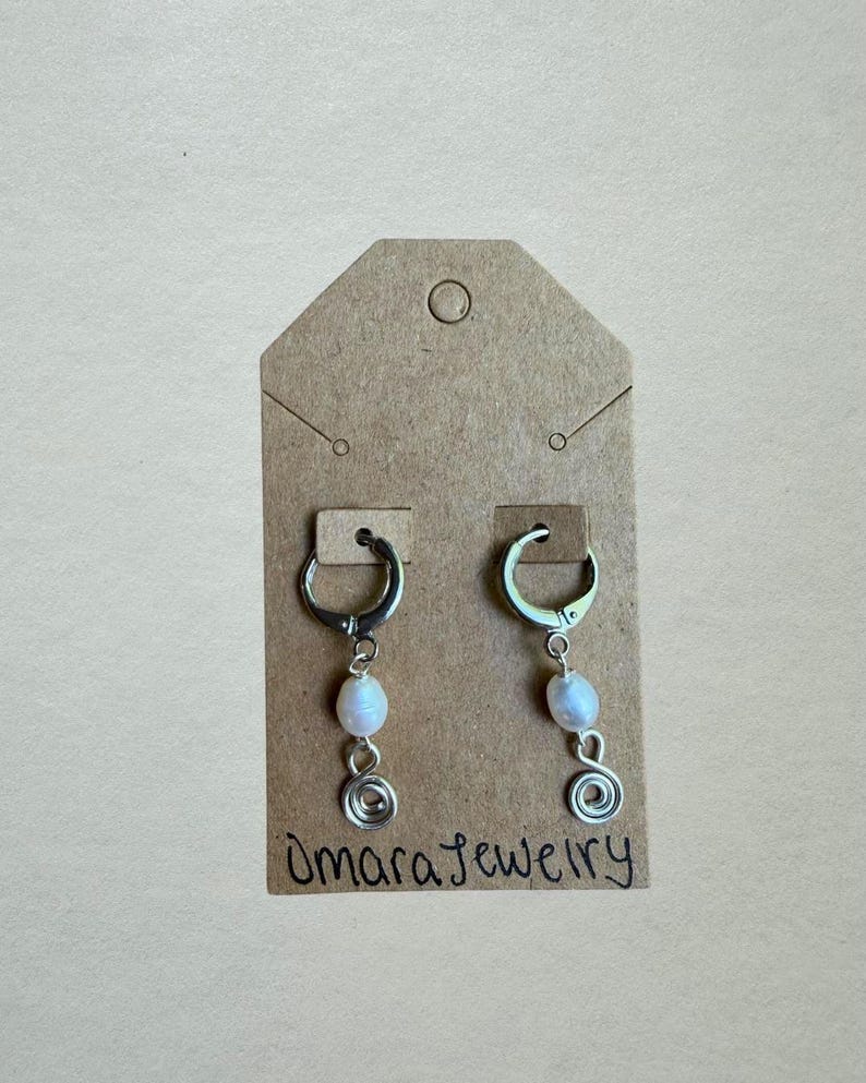 Puede incluir: Un par de pendientes de aro plateados con una cuenta de perla ovalada blanca y un dije de espiral de alambre plateado. Los pendientes est&aacute;n sujetos a una tarjeta de presentaci&oacute;n de cart&oacute;n marr&oacute;n con el texto Omara Jewelry escrito a mano.