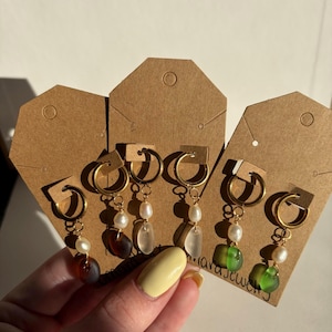 May include: Gold hoop earrings with dangling beads and pearls. The earrings feature a variety of colors, including amber, clear, and green. Each earring is attached to a brown tag.