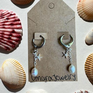 May include: A pair of silver hoop earrings attached to a brown cardboard display card labeled Umara Jewelry. Each earring features a small silver starfish charm and a dangling white freshwater pearl. The card is surrounded by various seashells.