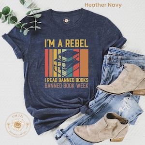 Könnte beinhalten: Marineblaues T-Shirt mit dem Text "I'M A REBEL" und "I READ BANNED BOOKS BANNED BOOK WEEK". Das Design zeigt einen Stapel Bücher. Das Shirt wird mit blauen Jeans und beigen Stiefeletten kombiniert.