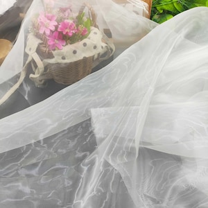 May include: Delicate white fabric with a gentle wave design, arranged on a surface. A wicker basket filled with pink flowers and a polka-dot lining is in the background, hinting at a decorative or craft use.