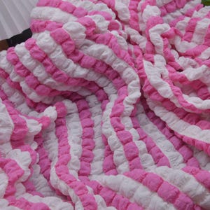 May include: A close-up of a soft, textured fabric with alternating vertical stripes of white and bright pink. The fabric appears to be gathered or ruched, creating a three-dimensional effect. The overall look is clean and modern.