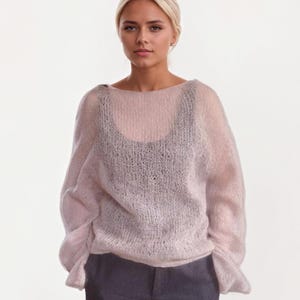 Puede incluir: Un suéter de punto rosa claro y transparente con mangas largas. El suéter se usa sobre una camiseta sin mangas negra y se combina con pantalones grises oscuros. El suéter tiene cuello barco y un ajuste holgado y fluido.